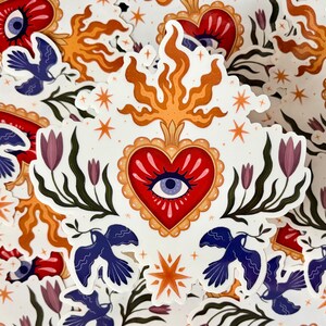 May include: A die-cut sticker featuring a central red heart with a blue eye at its center, surrounded by a gold scalloped border. Above the heart are orange flames and stars. Flanking the heart are green stems with purple tulips and blue birds.