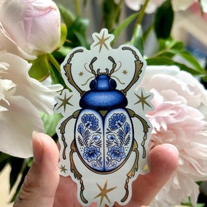 Op de afbeelding: Een sticker met een decoratief keverontwerp in blauw en wit met bloemmotieven. De kever heeft een blauwe kop en een witte buik met blauwe bloemmotieven. Gouden accenten en sterren omringen de kever. De sticker wordt voor roze en witte bloemen gehouden.