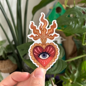 May include: A red heart with a gold frame and a single blue eye in the center. The heart is topped with a golden flame.