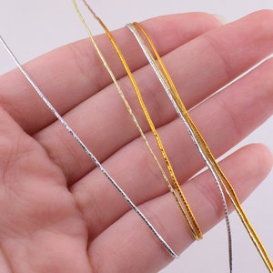 May include: Four strands of metallic cord, two gold and two silver, are held in a hand. The cords are thin and have a subtle shimmer.