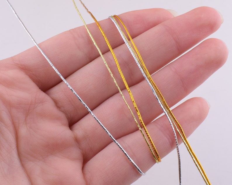 May include: Four strands of metallic cord, two gold and two silver, are held in a hand. The cords are thin and have a subtle shimmer.
