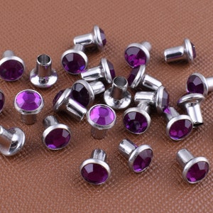 Peut inclure: Une collection de clous en strass violets avec des montures argentées. Les clous sont disposés de manière aléatoire sur une surface brune.