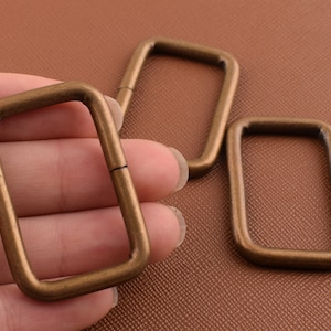Può includere: Tre anelli metallici rettangolari color bronzo antico. Un anello è tenuto in mano, mentre altri due sono posizionati vicino. Gli anelli hanno una forma rettangolare con angoli arrotondati e una fessura su un lato.