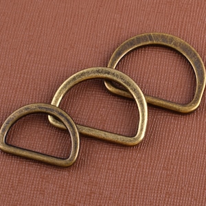 Op de afbeelding: Drie antieke goudkleurige metalen D-ringen in verschillende maten. De ringen zijn gestapeld, met de grootste ring onderaan en de kleinste ring bovenaan.