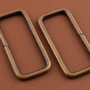 May include: Two antique gold-toned metal rectangular D-rings. The rings are smooth and have a slightly rounded edge.