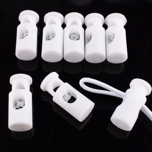 Puede incluir: Cierres de cordón de plástico blanco con un mecanismo de resorte. Los cierres de cordón están diseñados para asegurar cordones y cuerdas.