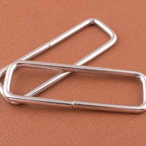 Puede incluir: Dos anillos en D de metal plateado, uno ligeramente más grande que el otro, están apilados uno encima del otro. Los anillos son lisos y tienen un acabado pulido.