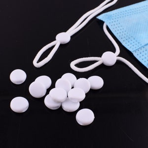 May include: White plastic mask strap adjusters, also known as ear savers, are scattered on a black surface. A white elastic cord with white plastic adjusters is also shown.