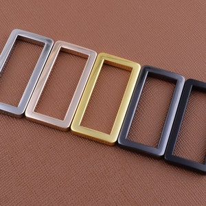 May include: Four metal rectangular rings in different finishes: silver, gold, black, and dark gray. The rings are arranged in a row from left to right, with the silver ring on the left and the dark gray ring on the right.