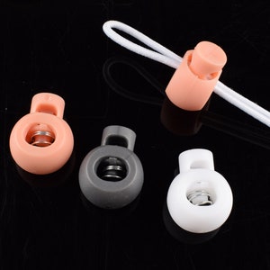 May include: Four toggle cord locks in different colors: pink, gray, white, and pink. Each lock has a spring inside and a hole for a cord to pass through.
