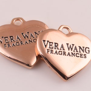 May include: Two rose gold heart-shaped charms with the text "Vera Wang Fragrances" engraved on them.
