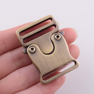 May include: A brass-colored metal buckle with a side release clasp. The buckle has a brushed finish and a slightly rounded shape.