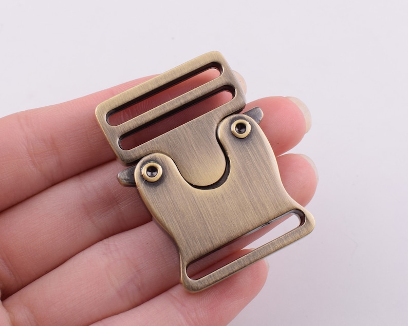 May include: A brass-colored metal buckle with a side release clasp. The buckle has a brushed finish and a slightly rounded shape.