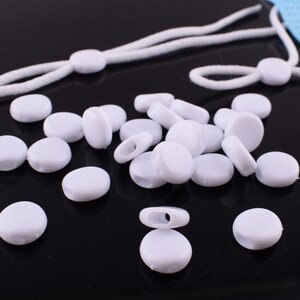 May include: A close-up of a pile of white plastic mask extenders. The extenders are round and have a small hole in the center. They are designed to be used with face masks to help adjust the length of the ear loops.