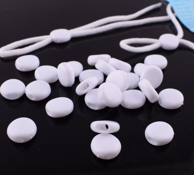 May include: A close-up of a pile of white plastic mask extenders. The extenders are round and have a small hole in the center. They are designed to be used with face masks to help adjust the length of the ear loops.