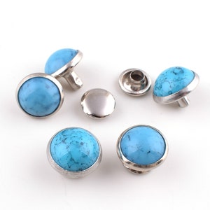 May include: Six turquoise blue and silver metal decorative studs. The studs have a round, flat top with a raised silver rim. The studs are designed to be used for clothing, bags, or other crafts.
