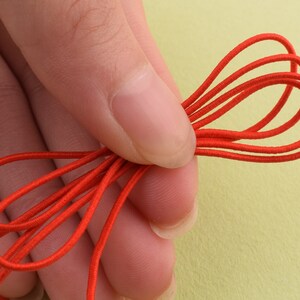 May include: Close-up of a bundle of bright red elastic cord. The cord is looped and held in a hand, showcasing its flexibility and color. The background is a soft yellow-green.