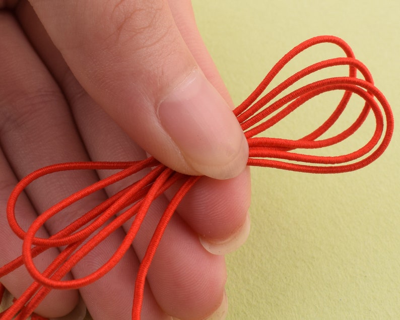 May include: Close-up of a bundle of bright red elastic cord. The cord is looped and held in a hand, showcasing its flexibility and color. The background is a soft yellow-green.