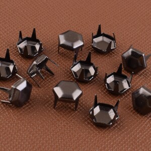 Puede incluir: Un grupo de 12 tachuelas hexagonales de metal negro con puntas puntiagudas. Las tachuelas están dispuestas en un patrón aleatorio sobre una superficie marrón.