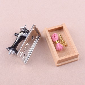 May include: A miniature sewing machine with a silver finish and black accents, next to a wooden box containing pink thread and gold scissors.
