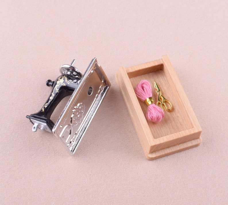 May include: A miniature sewing machine with a silver finish and black accents, next to a wooden box containing pink thread and gold scissors.