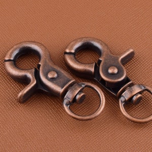 May include: Two antique copper-toned metal swivel snap hooks. The hooks have a spring-loaded gate and a D-ring.