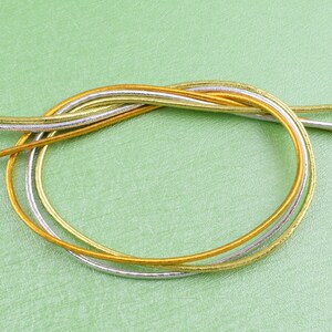 May include: Three strands of metallic cord in gold and silver colors are twisted together and laid out on a green surface.