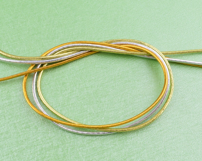 May include: Three strands of metallic cord in gold and silver colors are twisted together and laid out on a green surface.