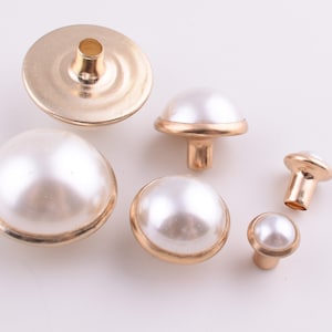 May include: Assortment of pearl buttons with gold metal accents. The buttons are in various sizes and shapes, including round, dome-shaped, and flat.