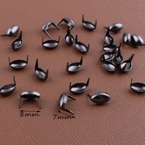 May include: A scattering of dark grey metal rivets, each with an oval head and two prongs. The rivets measure approximately 8 mm wide and 7 mm long. These decorative rivets are suitable for leatherwork, clothing, and various craft projects.