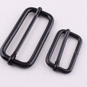 May include: Two black metal slider buckles, one larger than the other. The buckles are rectangular with rounded corners and have a smooth, shiny finish.