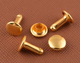 Può includere: Quattro rivetti in metallo color oro con testa rotonda e gambo cilindrico. I rivetti sono su una superficie marrone testurizzata.