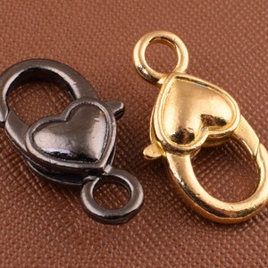 Könnte beinhalten: Zwei herzförmige Verschlüsse, einer goldfarben und einer schwarz, mit einer federbelasteten Öffnung. Diese Verschlüsse sind perfekt zum Anbringen an Schmuck, Taschen oder anderem Zubehör.