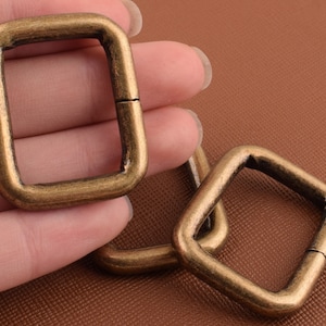 Op de afbeelding: Drie antieke goudkleurige metalen D-ringen. De ringen zijn vierkant met afgeronde hoeken en hebben een gladde afwerking.