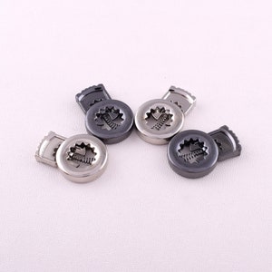 May include: A collection of metal buttons in various shades of gray and silver. Each button has a circular design with a detailed, stylized leaf pattern in the center. The buttons are scattered on a white surface.