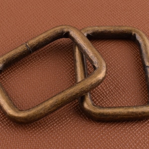 May include: Two antique gold-toned metal D-rings. The rings are square shaped and have a smooth, polished finish.