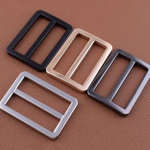 May include: Three metal slider buckles in different finishes: black, gold, and silver. The buckles are rectangular with two parallel bars.