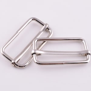 May include: Two silver metal slider buckles for straps or belts. The buckles are rectangular with rounded corners and have a smooth, polished finish.