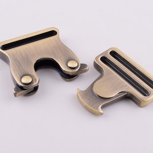 May include: Two antique gold metal clasps with black accents. The clasps are designed to be used for attaching straps or belts.