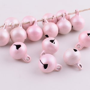 Peut inclure: Un groupe de clochettes de No&euml;l en m&eacute;tal rose avec une boucle argent&eacute;e pour accrocher. Les clochettes sont &eacute;parpill&eacute;es sur une surface blanche.