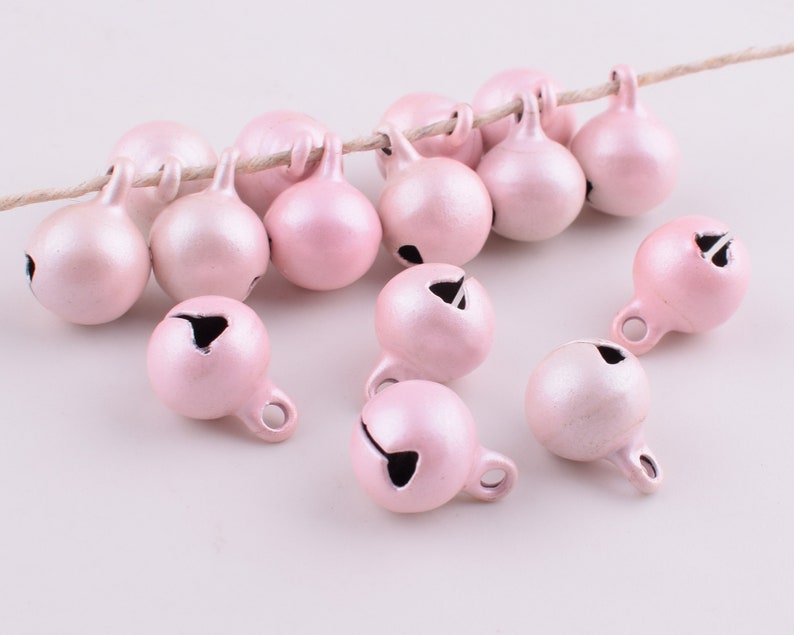Peut inclure: Un groupe de clochettes de No&euml;l en m&eacute;tal rose avec une boucle argent&eacute;e pour accrocher. Les clochettes sont &eacute;parpill&eacute;es sur une surface blanche.