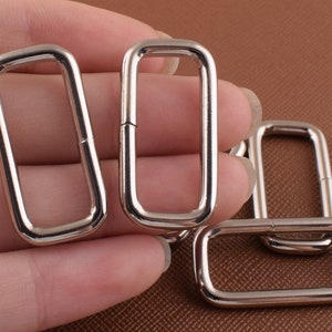 May include: Four silver metal rectangular rings, also known as D-rings, are shown in a close-up image. The rings are arranged in a pile, with one ring partially obscured by another. The rings are smooth and shiny, with a slightly rounded edge.