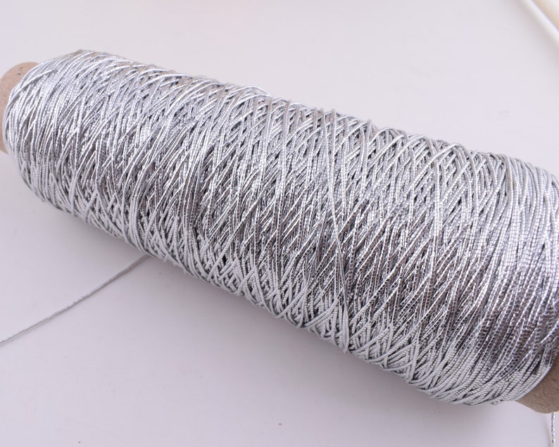 May include: A spool of silver metallic thread. The thread is wound tightly around the spool, creating a textured surface.