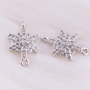 Könnte beinhalten: Zwei silberne Schneeflocken-Anhänger mit klaren Kristallverzierungen. Jeder Anhänger hat eine Öse oben und eine kleine Öse an der Seite.