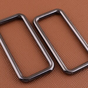 May include: Two black metal rectangular rings with rounded edges. The rings are lying on a brown textured surface.