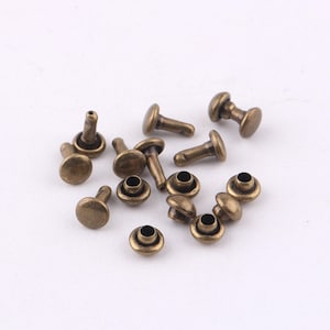 May include: A group of 14 antique gold-toned metal rivets. The rivets have a round head and a post that is designed to be hammered through a hole to secure two pieces of material together.