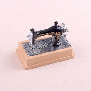 May include: A miniature black sewing machine with gold accents and a wooden base. The machine has a decorative floral design and a silver needle.