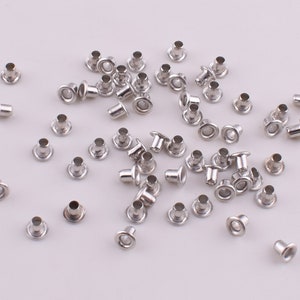 May include: A large quantity of silver metal eyelets, also known as grommets, are scattered on a white surface. These eyelets are used for reinforcing holes in fabric, leather, or other materials.