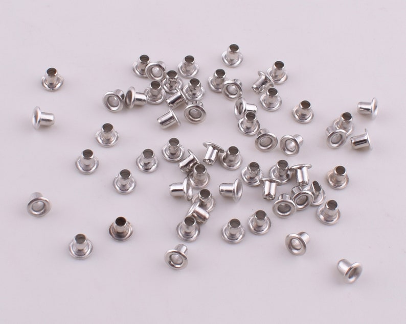 May include: A large quantity of silver metal eyelets, also known as grommets, are scattered on a white surface. These eyelets are used for reinforcing holes in fabric, leather, or other materials.