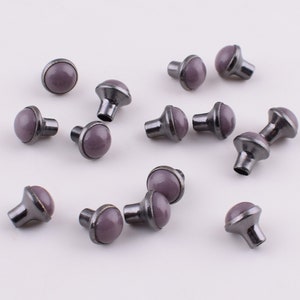Puede incluir: Una colección de 15 tachuelas de metal de color morado y negro con la parte superior redonda. Las tachuelas tienen un borde decorativo ligeramente elevado alrededor de la parte superior.